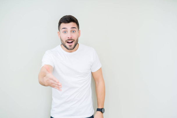 Mladý pohledný muž nosí ležérní bílé tričko izolované pozadí s úsměvem přátelskou nabídku handshake jako pozdrav a vítání. Úspěšné podnikání. - Fotografie, Obrázek