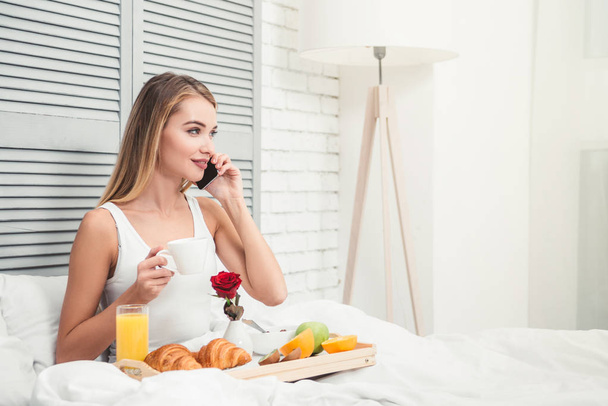 portrait of woman in the bed talking on smartphone while having breakfast - Foto, Imagem