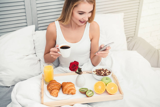 overhead view of young woman using smarthone while drinking coffee in the bed at the morning - Foto, afbeelding