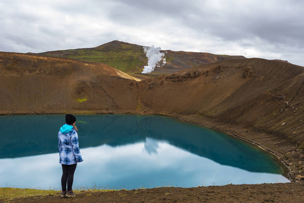 Girl teenager is staying in the border of Krafla volcanic crater and Viti lake. Vapor of geothermal power plant unit is at background, Nordurland eystra region in Northern Iceland. - Photo, Image