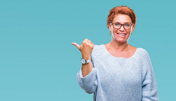 Atrractive senior caucasian redhead woman wearing glasses over isolated background smiling with happy face looking and pointing to the side with thumb up. - Photo, Image