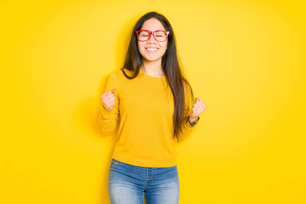 Beautiful brunette woman wearing red glasses over yellow isolated background very happy and excited doing winner gesture with arms raised, smiling and screaming for success. Celebration concept. - Valokuva, kuva