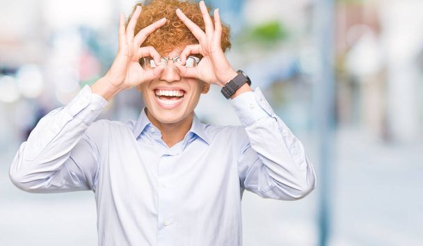 Mladý pohledný obchodní muž s afro brýle dělají ok gesto jako dalekohled držet jazyk, oči dívá skrze prsty. Šílený výraz. - Fotografie, Obrázek