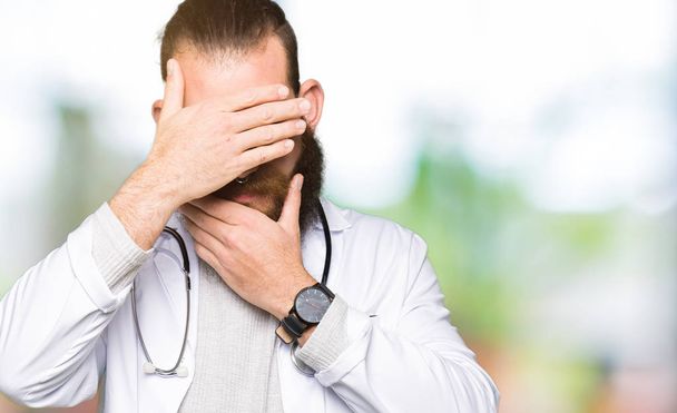 Mladá blonďatá lékař muž s plnovousem na sobě lékařský plášť pokrývající oči a ústa s rukama, překvapen a šokován. Skrývání emocí - Fotografie, Obrázek