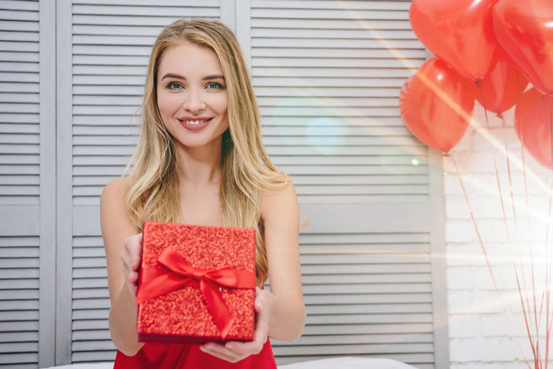 cheerful woman holding red gift box sitting on the bed and looking at the camera, valentines day concept - Fotoğraf, Görsel