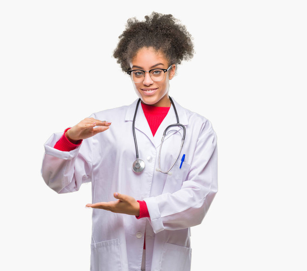 Young afro american doctor woman over isolated background gesturing with hands showing big and large size sign, measure symbol. Smiling looking at the camera. Measuring concept. - Foto, Imagen