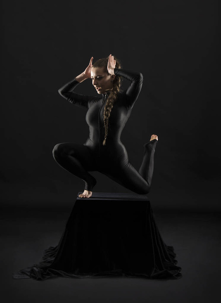 Girl in a black suit with long hair, performs gymnastic exercises. Studio shot on dark background. - Fotó, kép