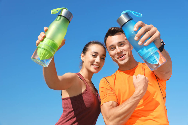 Young sporty couple holding bottles of water against blue sky on sunny day - Фото, зображення