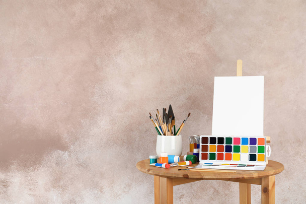 Wooden easel with blank canvas board and painting tools for children on table near color wall. Space for text - Foto, imagen
