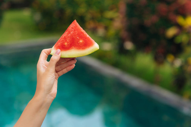 Close up slice of watermelon in female hand on background of blue swimming pool and greenery - Fotó, kép