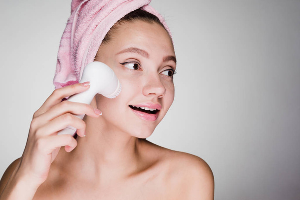 uma mulher com uma toalha na cabeça está realizando uma limpeza profunda do rosto com uma escova elétrica
 - Foto, Imagem