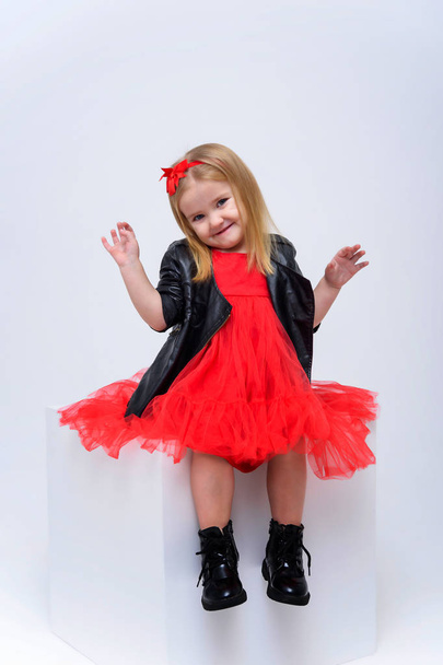 Concept portrait of a cute beautiful baby girl on a white background in a fashionable red dress. She sits on a cube right in front of the camera in various poses and shows different emotions. - Foto, Imagem