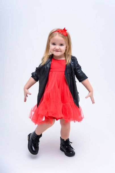Concept portrait of a cute beautiful baby girl on a white background in a fashionable red dress. She stands directly in front of the camera in different poses and shows different emotions. - Valokuva, kuva