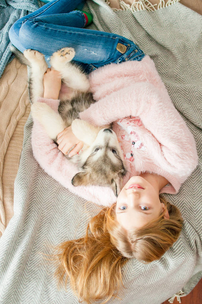 Cute girl with blond long hair plays with small Malamute puppy at home in a room decorated for Christmas - Fotó, kép