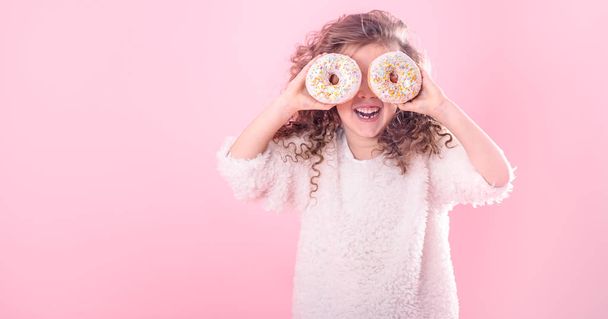 Portrét usměvavá holčička s kudrnatými vlasy a dvě chutné koblihy v dlaních, zavře oči s koblihy, na růžovém pozadí, místo pro text - Fotografie, Obrázek