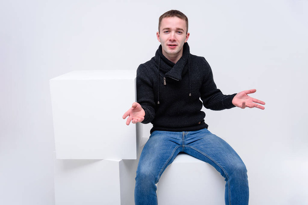 Concept studio portrait of a young man on a white background sitting on a cube and talking. He sits opposite the camera in different poses and with different emotions in jeans and a sweater. - Φωτογραφία, εικόνα