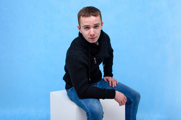 Concept studio portrait of a young man on a blue background sitting on a cube. He sits opposite the camera in different poses and with different emotions in jeans and a sweater. - Foto, imagen