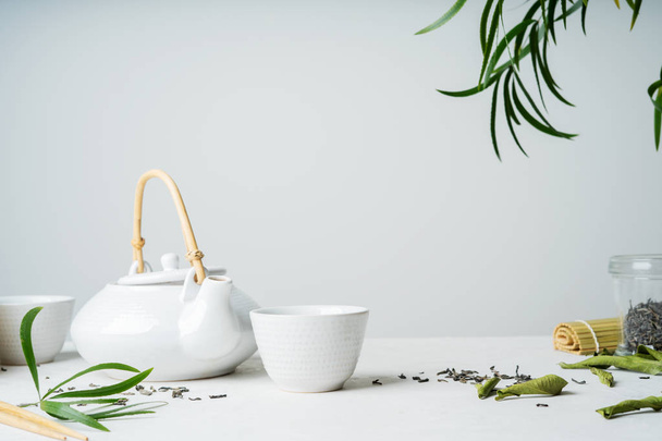 Asian tea set with two white cups of tea and teapot surrounded with green tea dry leaves on light grey background - Foto, Imagem