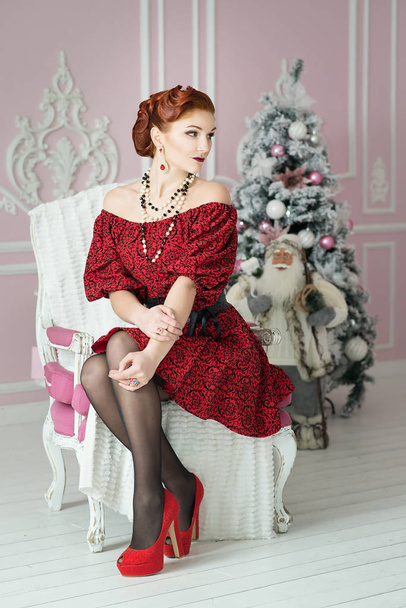 ヴィンテージの髪型とヴィンテージの椅子に座っている赤いドレスのメイクアップの豪華な若い女性 - 写真・画像