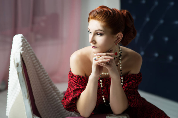 wunderschöne junge Frau mit Vintage-Frisur und Make-up in rotem Kleid sitzt auf Vintage-Stuhl - Foto, Bild