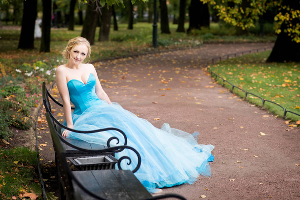 Blonde woman in sky blue fairy dress sitting on metal bench in autumn park - Фото, изображение