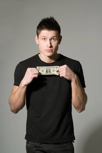 Portrait of young man in black t-shirt holding dollar on grey background - Foto, imagen