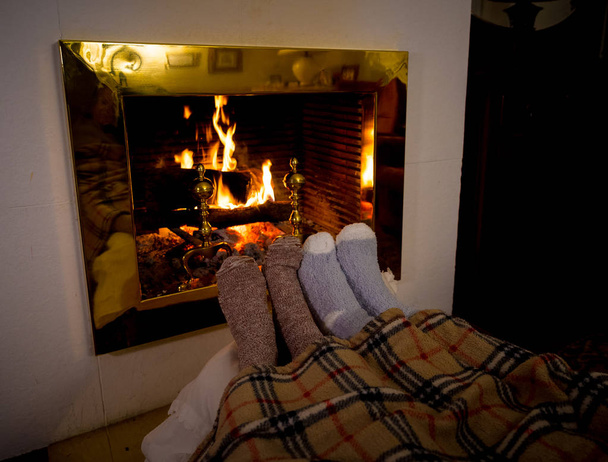 Imagen cercana de pareja sentada debajo de la manta junto a una acogedora chimenea calentando los pies en calcetines de lana relajándose con una taza de bebida caliente en casa en las frías vacaciones de invierno, momentos felices y navidad
. - Foto, imagen