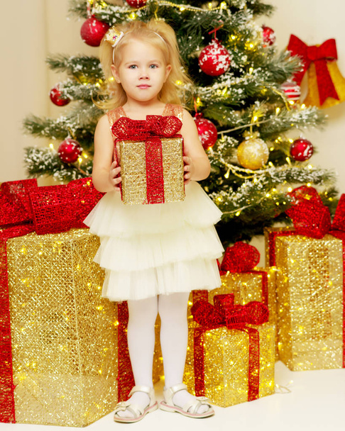 Little girl near the Christmas tree with a gift. - Фото, изображение