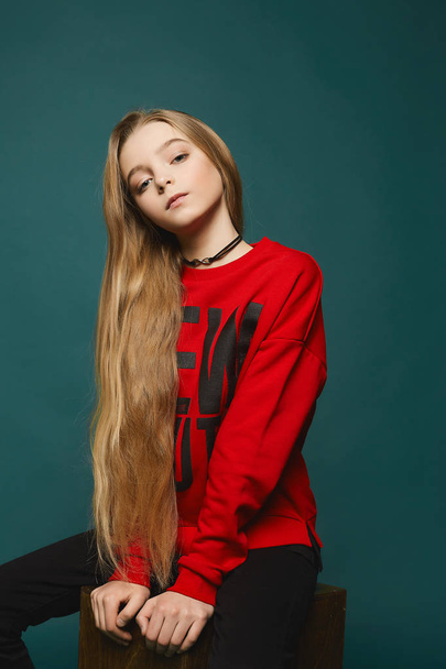 Stylish teen, beautiful young model girl with long blonde hair in black jeans and in fashionable red sweatshirt posing at the studio - Фото, зображення