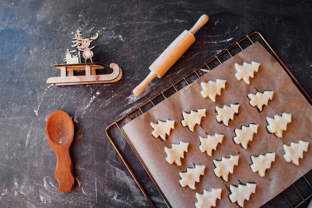 Raw dough cookies in Christmas tree shape ready to bake. Christmas Gingerbread cookies with baking paper on the tray on the dark background with wooden rolling pin and deer - Photo, Image