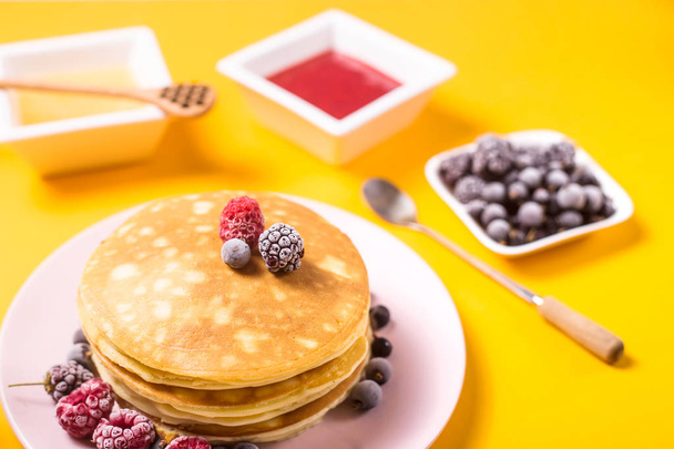 A stack of pancakes on a pink plate with berries next to honey and strawberry jam on a bright yellow background - Foto, Imagem