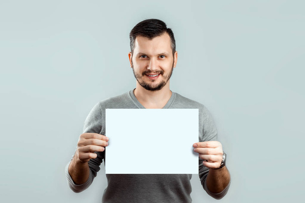 A young, attractive man holding a blank white A4 sheet, on a light background. mockup, layout, copy space. - Zdjęcie, obraz