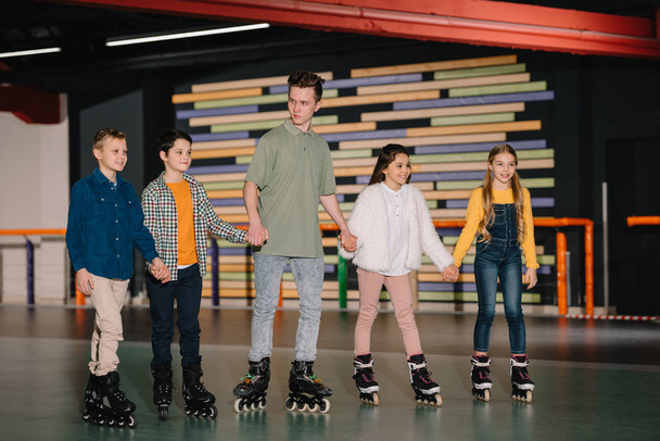 Pretty smiling children with handsome trainer standing in roller rink and holding hands - Foto, Imagem