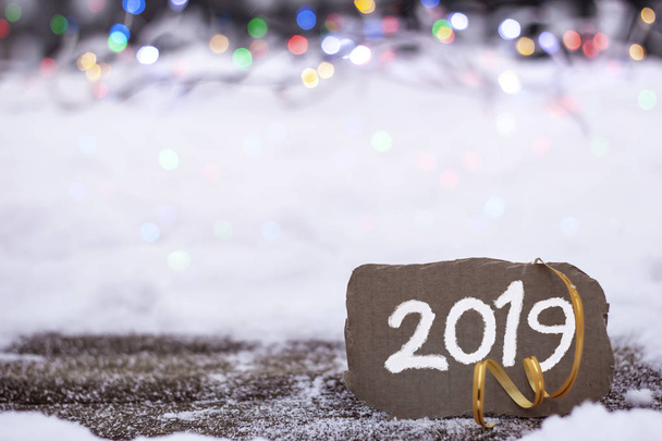 Бурый кусок картона с надписью 2019 года на фоне снега и ярких огней
 - Фото, изображение