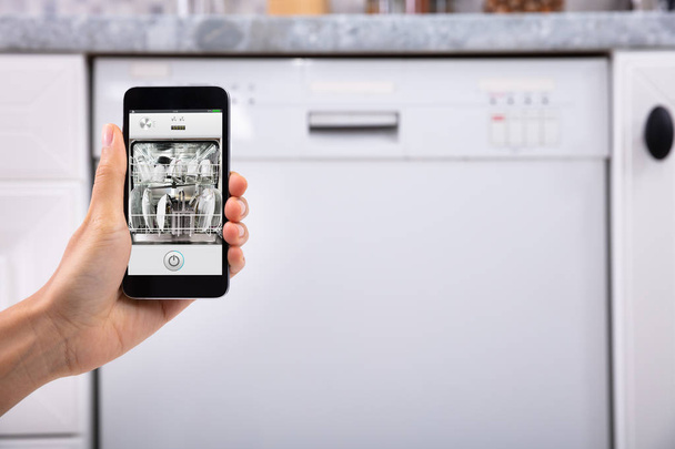Close-up Of A Woman's Hand Operating Dishwasher With Mobile Phone - Foto, imagen