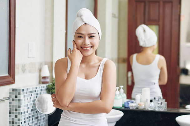 Nätti aasialainen nainen pyyhkeessä koskettaa pehmeää kasvojen ihoa ja hymyilee kameralle seisomassa kylpyhuoneessa suihkun jälkeen
 - Valokuva, kuva