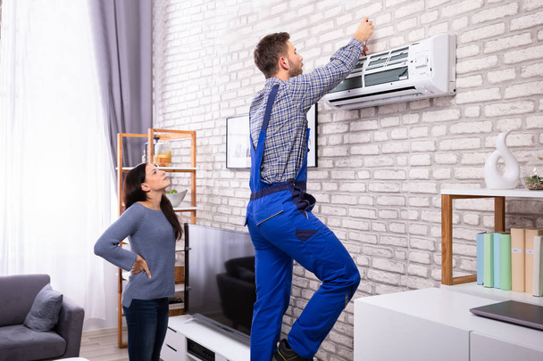 Young Woman Looking At Male Technician Repairing Air Conditioner Mounted On Brick Wall - Fotoğraf, Görsel
