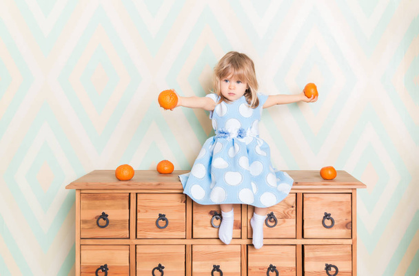 Toddler child baby girl in light blue dress seriously sitting on the wooden chest of drawers and holding fresh orange mandarins in her hands on rhomb wallpaper background. Children's emotions feelings. - Photo, image