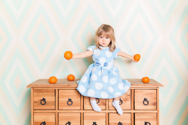 Toddler child baby girl in light blue dress seriously sitting on the wooden chest of drawers and holding fresh orange mandarins in her hands on rhomb wallpaper background. Children's emotions feelings. - Fotoğraf, Görsel