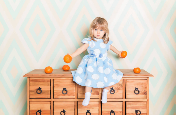 Toddler child baby girl in light blue dress seriously sitting on the wooden chest of drawers and holding fresh orange mandarins in her hands on rhomb wallpaper background. Children's emotions feelings. - Fotoğraf, Görsel
