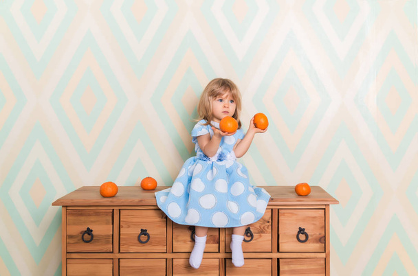 Toddler child baby girl in light blue dress seriously sitting on the wooden chest of drawers and holding fresh orange mandarins in her hands on rhomb wallpaper background. Winter holidays or christmas - Foto, Imagem