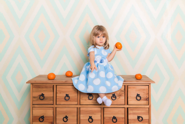 Little baby girl sitting cross-legged on chest of drawers holding tangerine in hand and pointing down with index finger on rhomb wallpaper background. Child in blue polka dot dress and white socks - Фото, изображение