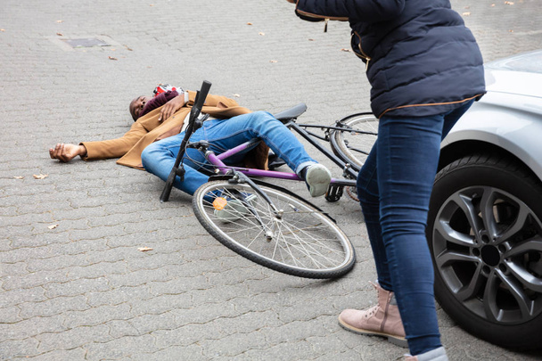 Young Woman Looking At Unconscious Male Cyclist Lying On Street After Accident Near Car - Foto, Imagen