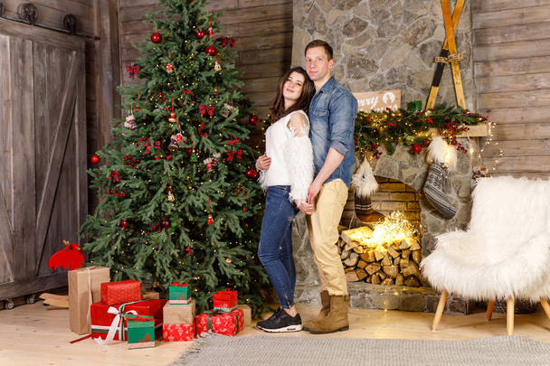 Young beautiful couple is standing near the fireplace on the background of the Christmas tree and smiling - Valokuva, kuva