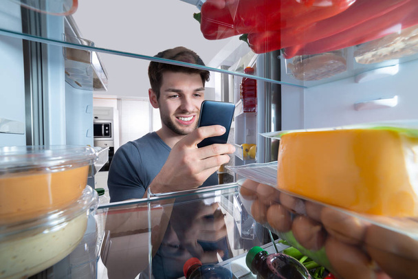 Happy Man Standing In Front Of Refrigerator Using Mobilephone In Kitchen - Foto, immagini