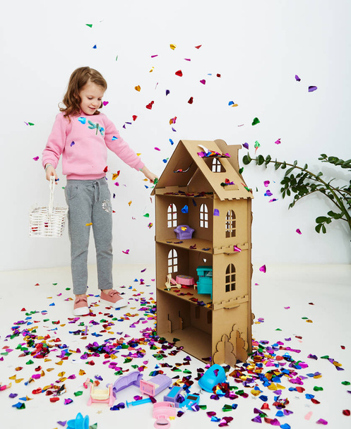 Happy beautiful little girl enjoying colorful confetti surprise falling down, posing on white studio wall. Pretty girl celebrating her birthday party, having fun - Φωτογραφία, εικόνα
