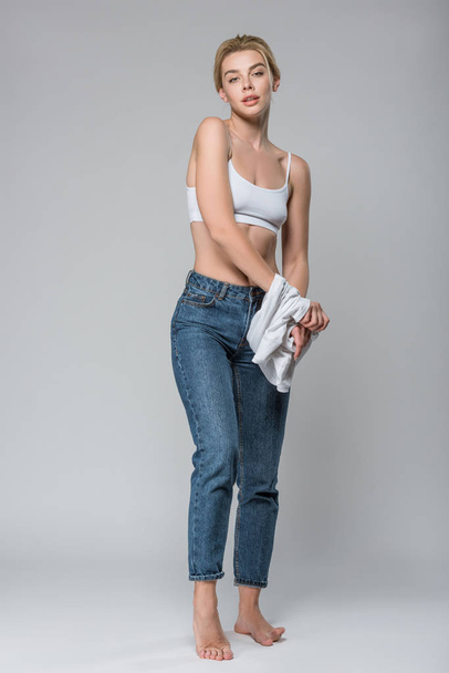 slim blonde girl in jeans and white bra undressing isolated on grey - Фото, изображение