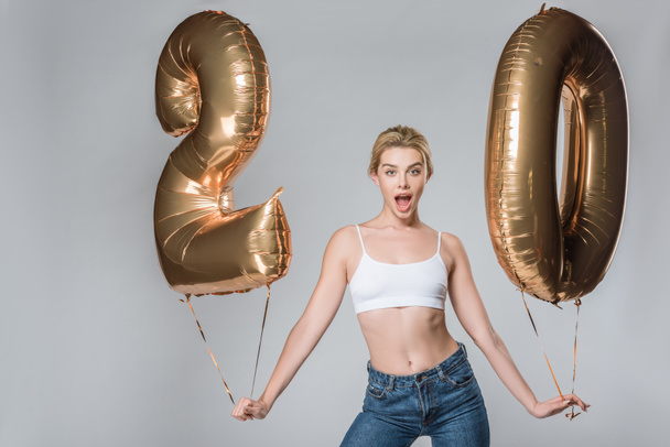 surprised beautiful girl in jeans and white bra posing with 20 golden balloons, isolated on grey - Zdjęcie, obraz