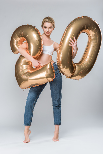 attractive girl in jeans and white bra posing with 20 golden balloons, on grey - Фото, зображення