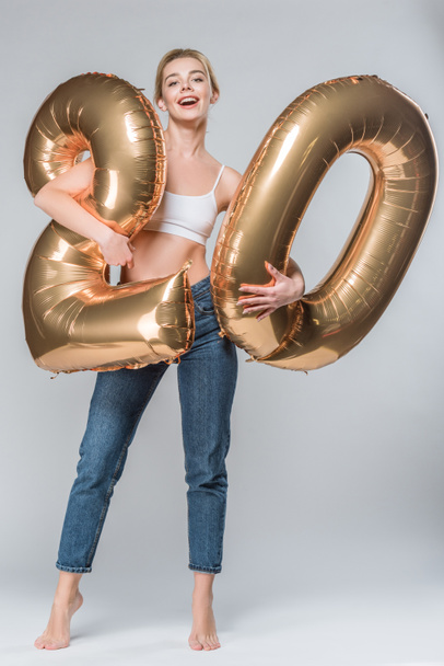 happy girl in jeans and white bra posing with 20 golden balloons, on grey - Foto, imagen